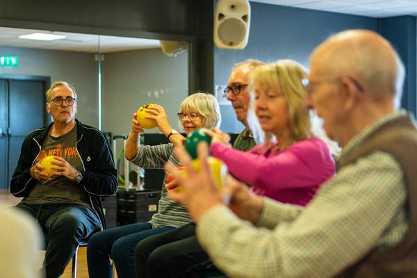 Quelle: Centre for Ageing Better ine Gruppe älterer Menschen sitzt im Halbkreis auf Stühlen in einem hellen Raum und führt gemeinsam Bewegungsübungen mit bunten Gymnastikbällen durch. Alle wirken konzentriert und motiviert, einige lächeln dabei. Die Atmosphäre ist aktiv und gemeinschaftlich.